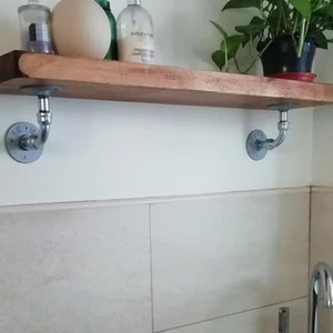 May include: A wooden shelf with metal pipe brackets, holding a plant, a bottle of lotion, and a decorative egg. The shelf is mounted on a white tiled wall.