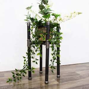 Puede incluir: Un soporte para plantas de metal negro con cuatro patas, que sostiene una planta en maceta con flores blancas y hojas verdes. El soporte tiene un diseño sencillo y está hecho de tubería industrial.