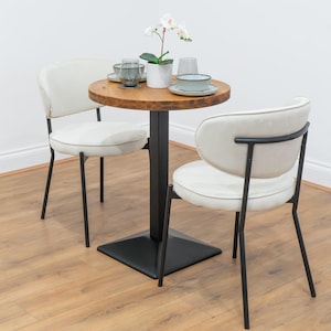 May include: A compact dining set featuring a round wooden table and two cream-coloured chairs. The table has a black metal base and is set with tableware and a potted orchid. The chairs have black metal frames and padded seats.