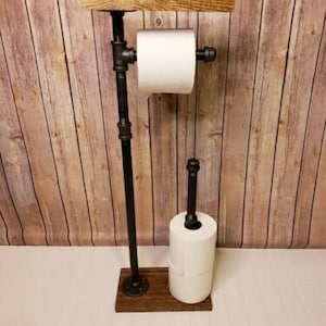 May include: A rustic toilet paper holder made of black metal pipes and a wooden shelf. The holder has two rolls of toilet paper, one on top of the other.