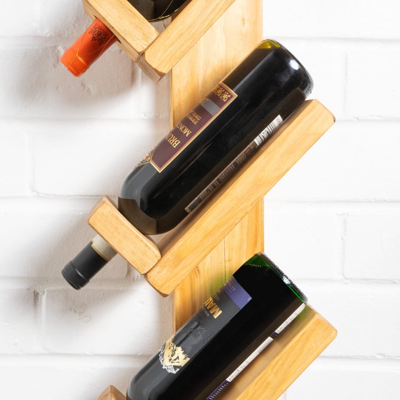 Op de afbeelding: Een houten wandrek voor wijn met een trapsgewijs ontwerp. Het rek heeft ruimte voor vijf flessen wijn, waarvan er drie zichtbaar zijn op de afbeelding. De flessen zijn een mix van rode en witte wijn.