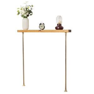 Può includere: Una mensola in legno con gambe in ottone, con un vaso bianco con fiori, una sveglia nera e una lampada marrone scuro con copertura in vetro. La mensola è in legno chiaro.