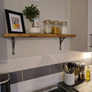 Puede incluir: Una estanter&iacute;a de madera con soportes met&aacute;licos, que sostiene una peque&ntilde;a planta en maceta, una impresi&oacute;n enmarcada con el texto "The Kit" y tres tarros de vidrio llenos de pasta y harina. La estanter&iacute;a est&aacute; montada en una pared de azulejos blancos.