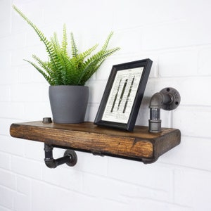 Op de afbeelding: Een rustiek houten plank met zwarte metalen pijpbeugels. De plank is versierd met een ingelijst drukwerk, een pot met varen en een zwarte metalen pijpfitting.