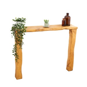 May include: A rustic wooden console table with a natural wood finish. The table has a single shelf and two legs that are shaped like tree trunks. There are two small glass bottles and a potted plant on the shelf.