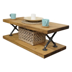 May include: A rustic wooden coffee table with a lower shelf. The table has black metal X-shaped legs and a woven basket on the lower shelf. Two teal mugs and a white candle are on the top shelf.