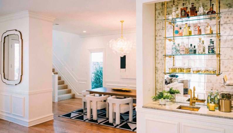 May include: A white dining room with a large glass chandelier, a wooden table with white chairs, and a mirrored bar with gold shelves and bottles.