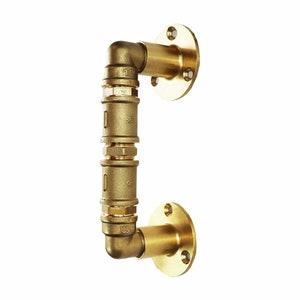 May include: Brass door handle with a pipe design. The handle is made of multiple sections of pipe connected together with fittings. The handle is attached to a wall with a round flange.