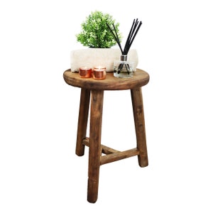 Könnte beinhalten: Ein Holzhocker mit runder Oberseite und drei Beinen. Der Hocker ist mit einem weißen Handtuch, zwei Kupferkerzen und einem Glasdiffusor mit schwarzen Schilfrohren verziert.
