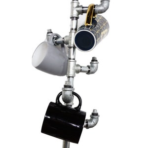 Pode incluir: Um suporte para canecas de metal feito de conex&otilde;es de tubo galvanizado. O suporte tem quatro bra&ccedil;os que seguram canecas de diferentes tamanhos e cores. O suporte &eacute; montado em uma base de madeira.