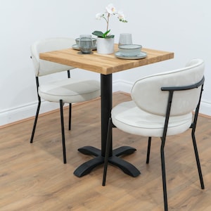 May include: A small dining set featuring a square wooden table and two cream-colored chairs. The table has a black pedestal base and is set with plates, glasses, and a potted orchid. The chairs have black metal frames.