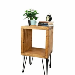 May include: A wooden side table with a square design and black hairpin legs. The tabletop holds a small potted plant, a light blue mug, a stack of books, and a black alarm clock. The table is made of light brown wood.