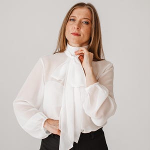 May include: A white blouse with a high collar and a large bow tie. The long-sleeved blouse has a sheer appearance and is paired with black pants. The overall style is elegant and suitable for formal or business attire.