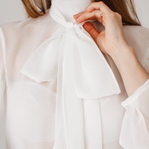 May include: A white, sheer blouse with a high neckline and a large bow detail. The long-sleeved top is made of a lightweight fabric, creating a flowing silhouette. The bow is tied at the neck, adding a touch of elegance to the garment.