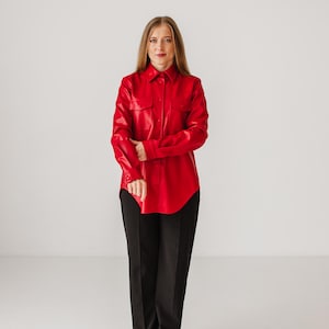 Puede incluir: Una camisa roja brillante con botones, dos bolsillos en el pecho y mangas largas. La camisa se combina con pantalones negros. El conjunto es un look audaz y monocromático, adecuado para una variedad de ocasiones.