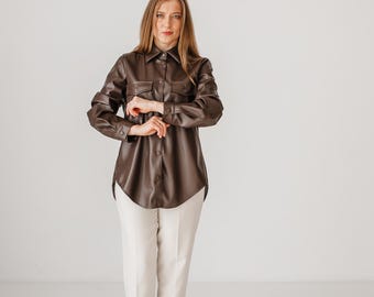 May include: A brown leather shirt with two chest pockets and a button-down front. The shirt is paired with cream-colored pants and black heels. The outfit is styled in a studio setting with a white background.