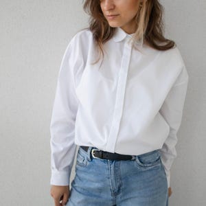 Peut inclure: Une chemise blanche à col rond et manches longues, associée à un jean bleu clair et une ceinture noire. L'ensemble est simple et classique.