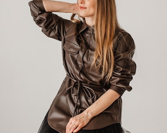 May include: A dark brown faux leather button-down shirt featuring long sleeves, a pointed collar, two chest pockets with flaps, and a matching tie belt at the waist, paired with a layered black tulle skirt with a sheer, voluminous texture.