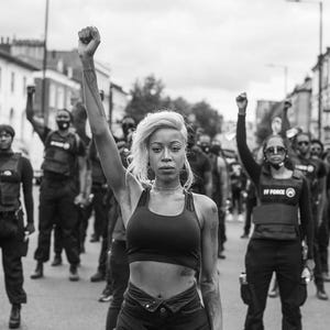 May include: A monochrome image of a demonstration. A group of people, many with raised fists, march in the street. The central figure is a person with blonde hair, wearing a black sports bra and dark trousers. Others wear black clothing and vests.