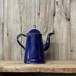 Könnte beinhalten: Eine Vintage-Kaffeekanne aus kobaltblauem Emaille mit Deckel und gebogenem Ausguss. Die Kaffeekanne steht auf einem rustikalen Holzregal vor einem verwitterten Holzhintergrund. Die Kanne hat einen Griff und einen kleinen Knopf auf dem Deckel.