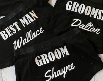 May include: A collection of black boxer briefs featuring white heat-pressed text. The garments display various titles and names including Groom Shayne, Best Man Wallace, Groomsman Dalton, and others labelled Saba and Connor in white lettering.