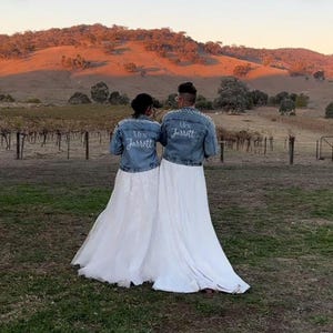 May include: Two people wearing white dresses and denim jackets with the text "Mrs. Jarrott" and "Mrs. Jarrott" embroidered on the back. They are standing in a field with a vineyard in the background.