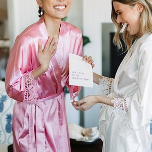 May include: Two women in silk robes, one pink and one white, are laughing. The pink robe has lace trim. One woman holds a card that reads "Will you be my bridesmaid?" A wedding proposal theme.