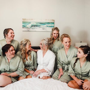 May include: A group of six women wearing matching sage green robes with lace trim. They are smiling and laughing together. One woman is wearing a white robe with lace trim.