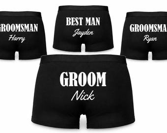 May include: Four pairs of black boxer briefs featuring white printed text. One pair reads GROOM Nick, one reads BEST MAN Jayden, and two pairs read GROOMSMAN Harry and GROOMSMAN Ryan. The text is centred on the front of each pair of underwear.