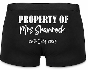 May include: Black boxer briefs featuring white text that reads PROPERTY OF Mrs Shamrock and the date 27th July 2026. The garment has a wide elastic waistband and a solid black fabric construction with clear white lettering printed on the centre.