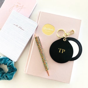 May include: A pink notebook with the words "Gratitude Journal" on the front. A black compact mirror with the initials "TP" is attached to a gold key chain. A pink pen with glitter is on the notebook.