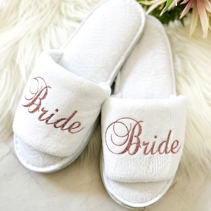 May include: Two white terry cloth slippers, each embroidered with the word "Bride" in a copper-coloured script. The open-toed slippers are displayed on a white, fluffy surface.