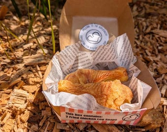 May include: A brown cardboard box containing two golden-brown fried chicken leg-shaped soaps wrapped in newspaper-print paper. The box features a sticker with a duck logo and text that reads: This box contains: 2 Fried Chicken Leg Soaps.