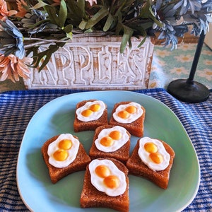 May include: A turquoise plate displays six pieces of toast, each topped with a fried egg. The toast is a golden brown, and the eggs have white and yellow details. A wooden planter with flowers is in the background.