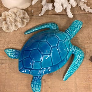 Puede incluir: Una figurita de tortuga de cerámica turquesa con una concha detallada y patas texturizadas. La tortuga está tumbada boca abajo con la cabeza girada hacia un lado.