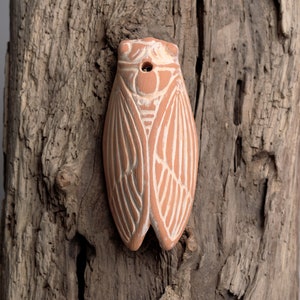 Może przedstawiać: Terakotowa rzeźba świerszcza z białymi detalami. Rzeźba jest wykonana z gliny i ma otwór na górze. Stoi na kawałku zwietrzałego drewna.