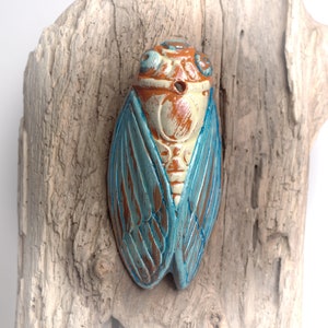 Può includere: Una statuetta di cicala in ceramica con ali blu e marroni e una testa bianca e arancione. La statuetta è montata su un pezzo di legno galleggiante.