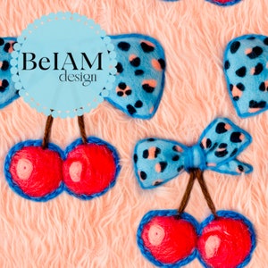 May include: Two felted cherry ornaments with bright red fuzzy spheres, brown stems, and light blue bows featuring a black and peach leopard print pattern. The text BeIAM design is printed in black on a light blue scalloped circular background.