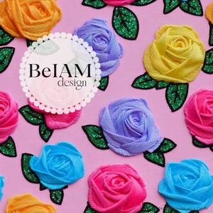 May include: A pink fabric background featuring a repeating pattern of embroidered roses in yellow, purple, blue, and pink, each accompanied by green glittery leaves. A circular white logo with the text BeIAM design is centred on the image.