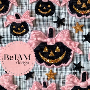 以下が含まれることがあります： 黒と金のハロウィンプンキンオーナメントにピンクのリボンが付いています。オーナメントは黒と白の市松模様の布地に置かれています。ピンクの円の中に「BeIAM design」の文字があります。