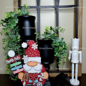 May include: A wooden Christmas gnome figurine with a red hat, white beard, and a red and white striped shirt. The gnome is holding a gingerbread cookie and a candy cane. The gnome is standing on a wooden base with the words "North Pole" written on it. The gnome is surrounded by greenery and black vases.