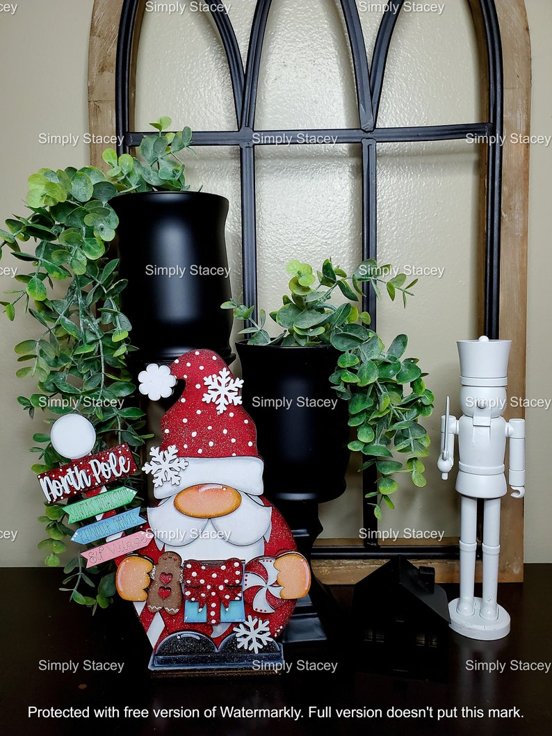 May include: A wooden Christmas gnome figurine with a red hat, white beard, and a red and white striped shirt. The gnome is holding a gingerbread cookie and a candy cane. The gnome is standing on a wooden base with the words "North Pole" written on it. The gnome is surrounded by greenery and black vases.