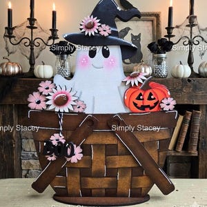May include: A wooden cutout decoration featuring a white ghost wearing a black witch hat with pink flowers, sitting in a brown woven basket. The ghost is accented with pink cheeks and accompanied by a bright orange jack-o'-lantern and the word BOO.