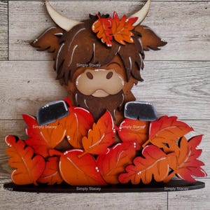 May include: A wooden cutout of a Highland cow with brown fur and a black nose, surrounded by orange and red fall leaves. The cow is wearing a crown of leaves.