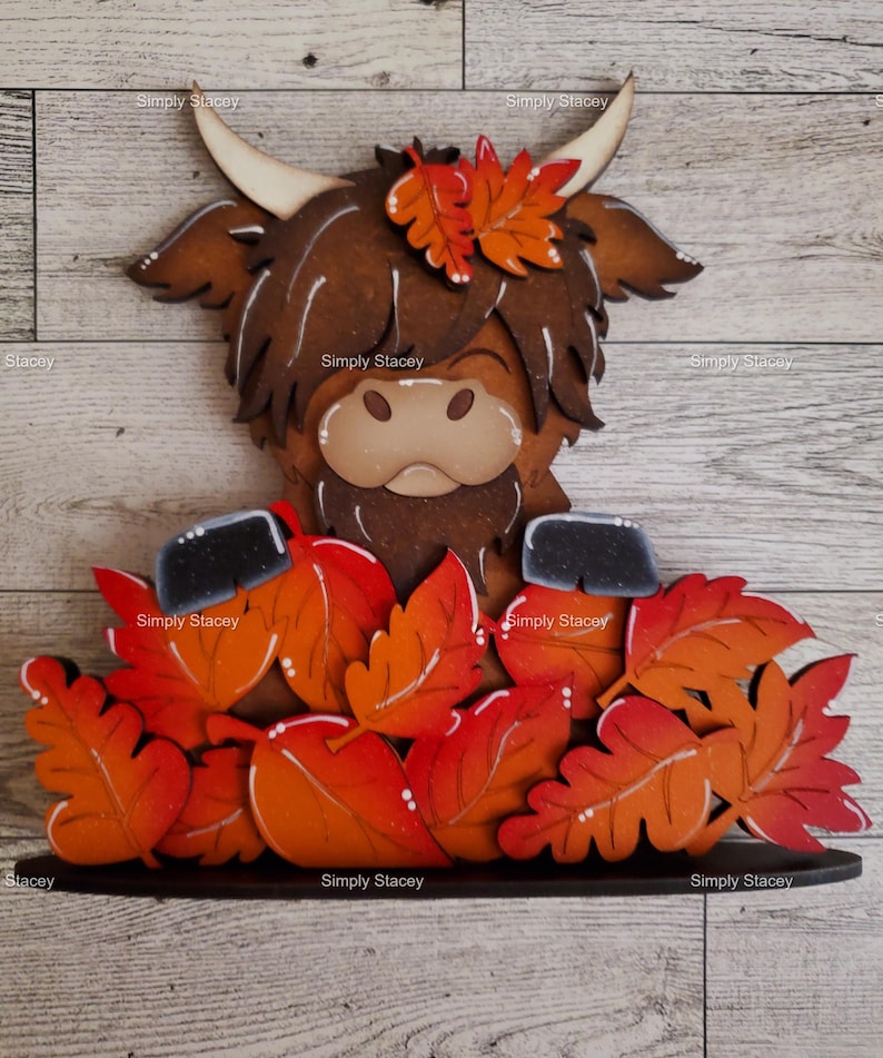 May include: A wooden cutout of a Highland cow with brown fur and a black nose, surrounded by orange and red fall leaves. The cow is wearing a crown of leaves.