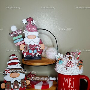 May include: Three Christmas gnomes, one with a red hat and white beard, one with a brown hat and white beard, and one with a red and white hat and brown beard. The gnomes are standing on wooden shelves. A red mug with white lettering that says "Mrs. Claus" is on the bottom shelf. The mug is filled with white whipped cream and topped with candy canes and gingerbread men.