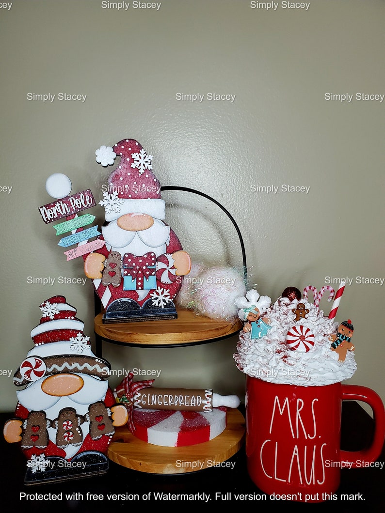 May include: Three Christmas gnomes, one with a red hat and white beard, one with a brown hat and white beard, and one with a red and white hat and brown beard. The gnomes are standing on wooden shelves. A red mug with white lettering that says "Mrs. Claus" is on the bottom shelf. The mug is filled with white whipped cream and topped with candy canes and gingerbread men.