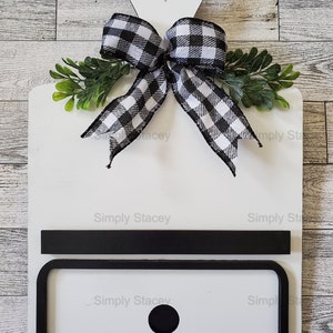 May include: White wooden cutting board with a black and white checkered bow, green leaves, and a black chalkboard insert. The board has a heart-shaped cutout at the top and the text "Simply Stacey" is written on the board.