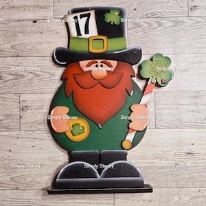 May include: A wooden leprechaun figurine with a green hat, red beard, and green shirt. He is holding a gold coin with a shamrock on it and a candy cane with a green shamrock at the top. The sign reads "Luck of the Irish".