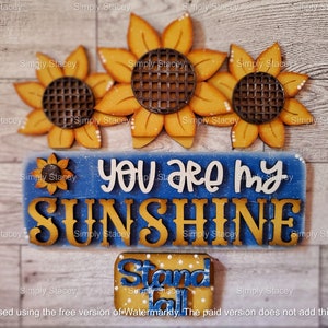 May include: A wooden sign with the text "You are my sunshine" in white letters on a blue background. Three yellow sunflowers with brown centers are above the sign. A smaller yellow sign with the text "Stand Tall" in blue letters is below the sign.